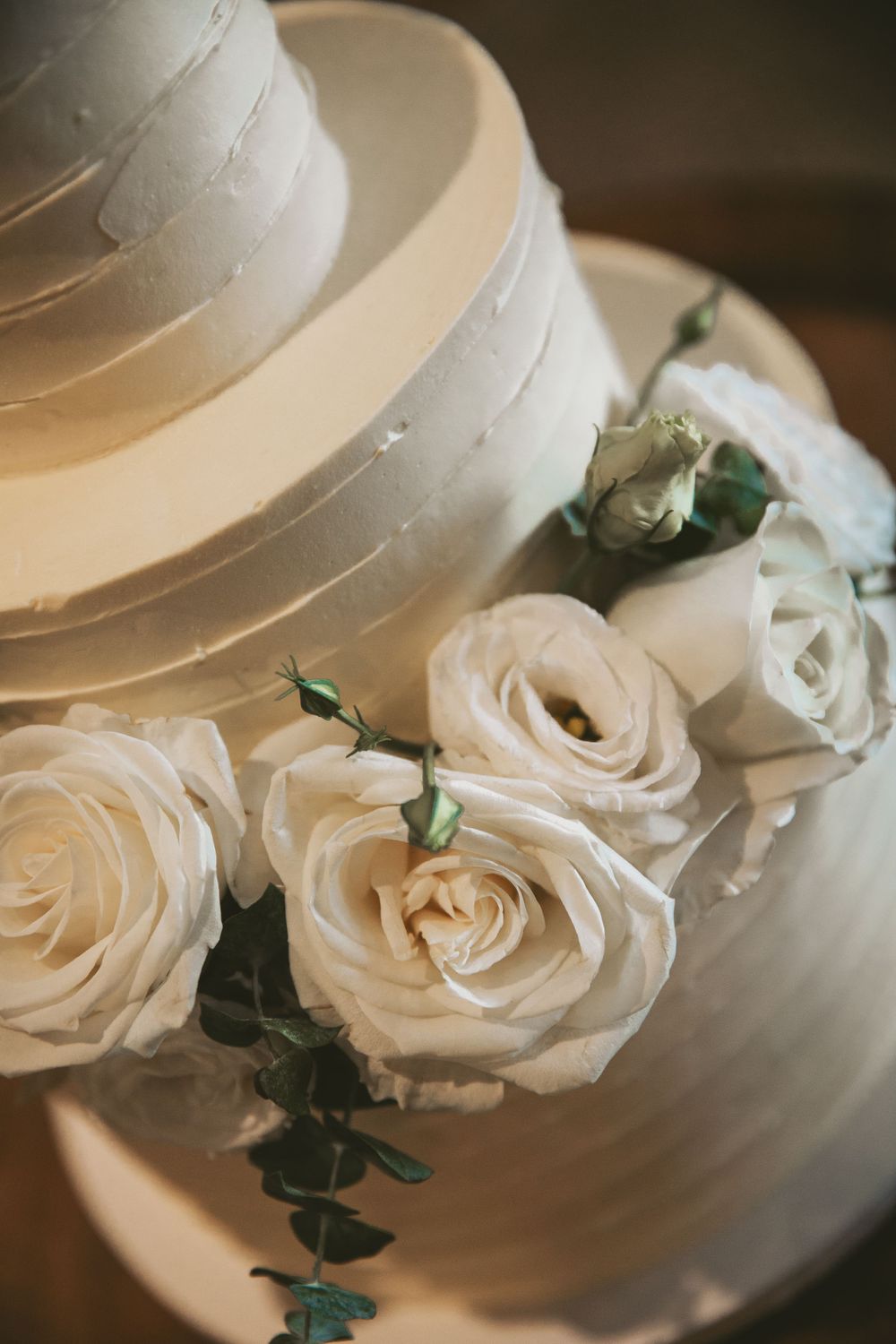 Close-up of white wedding cake tiers adorned with fresh roses and eucalyptus.