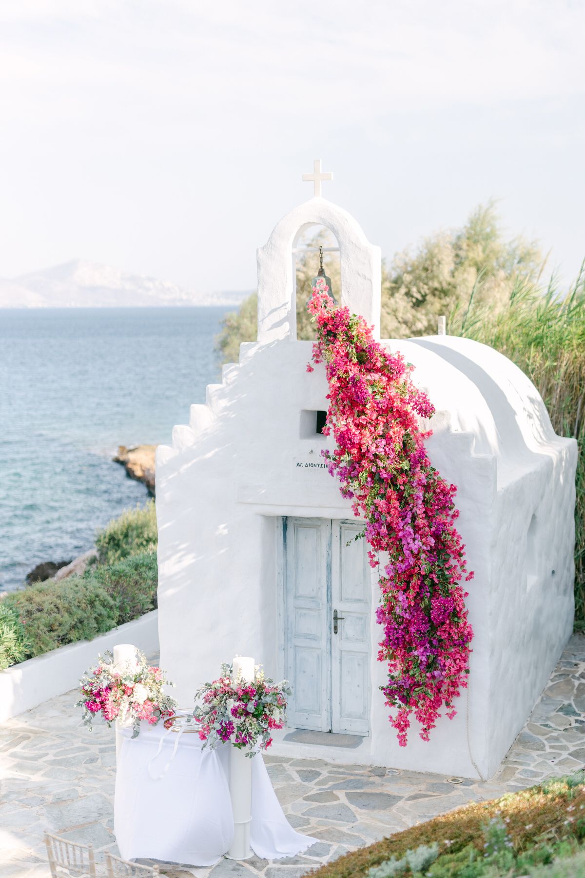 Small white Greek chapel with pink bougainvillea flowers by Mediterranean Sea on sunny day.