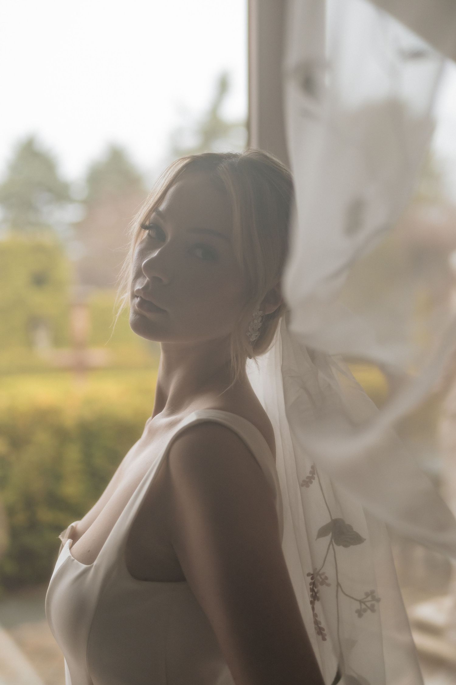 Ethereal bridal portrait backlit by soft natural light with flowing sheer veil in the breeze.