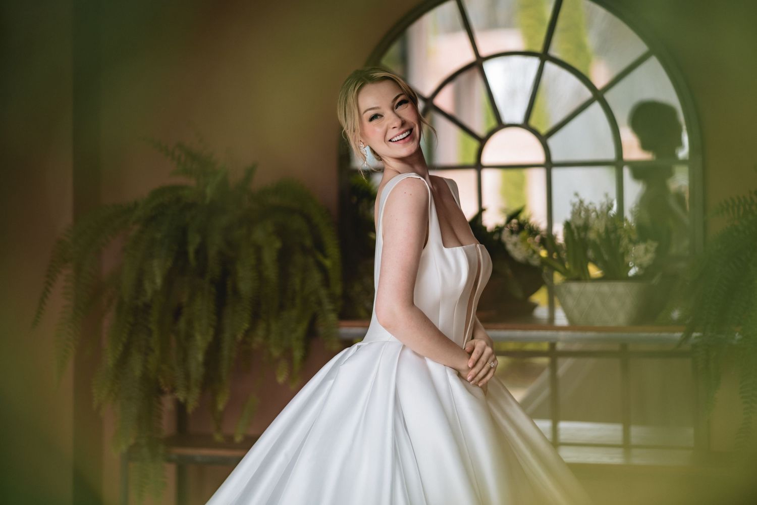 Elegant white wedding dress photographed in front of an arched window with potted ferns creating a romantic indoor setting.
