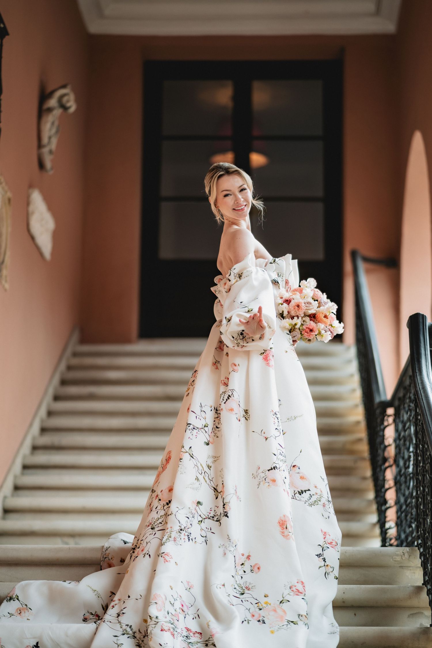 Elegant wedding gown with delicate floral pattern displayed on grand staircase with peach-colored walls.
