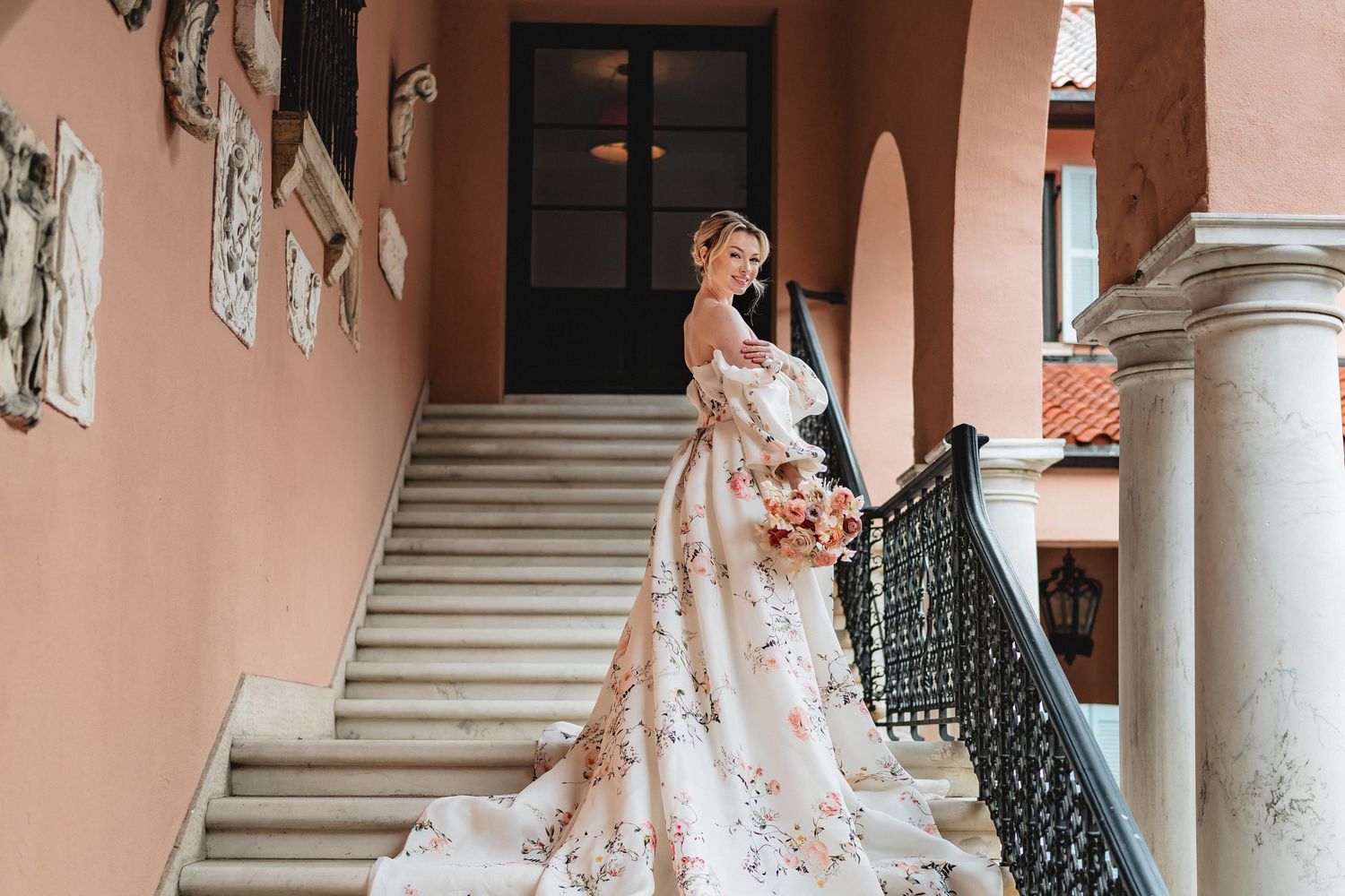 Elegant bride in floral lace gown with flowing train stands on grand staircase against peach-colored walls.