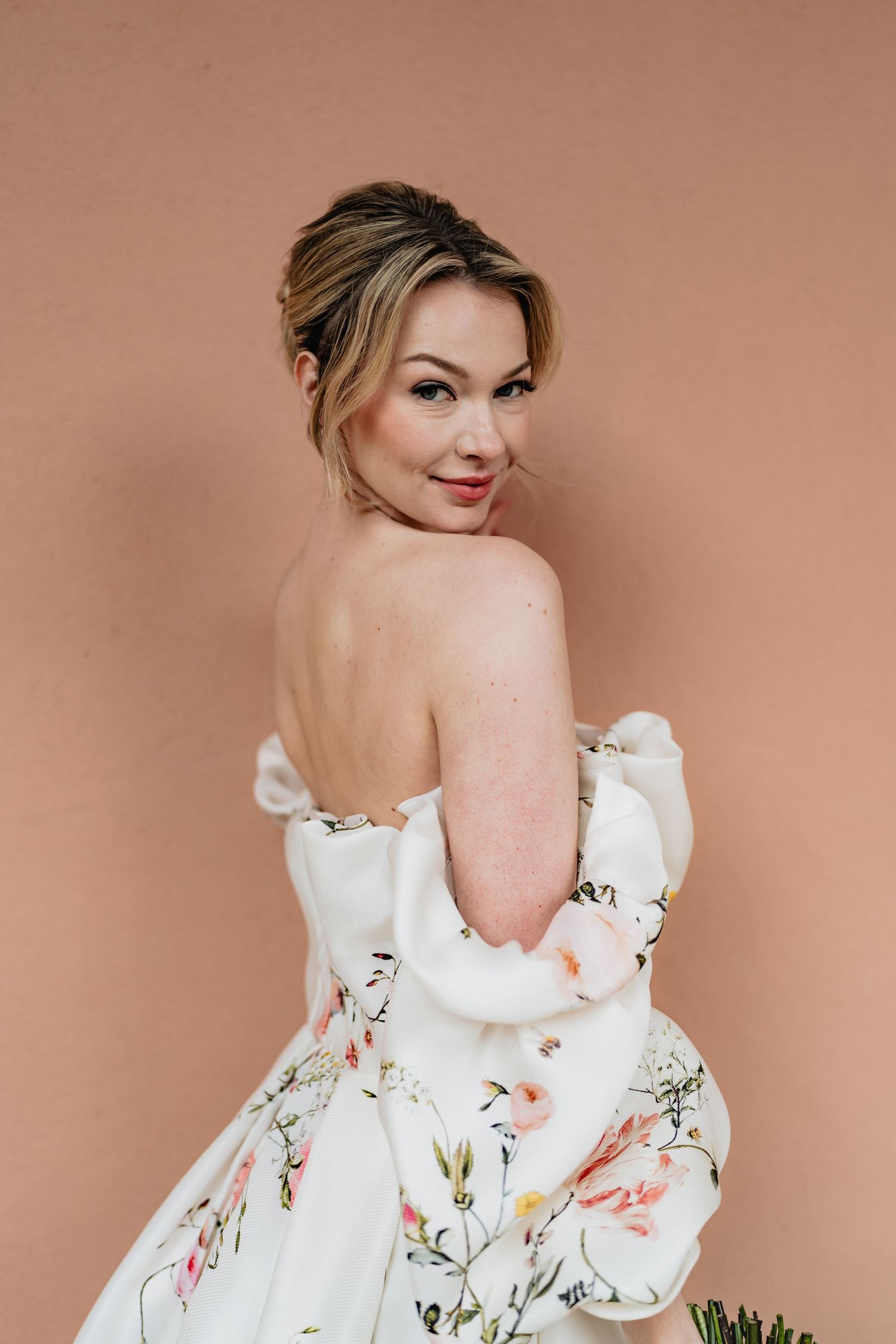Woman wearing white floral off-shoulder dress poses against beige background in romantic portrait style.