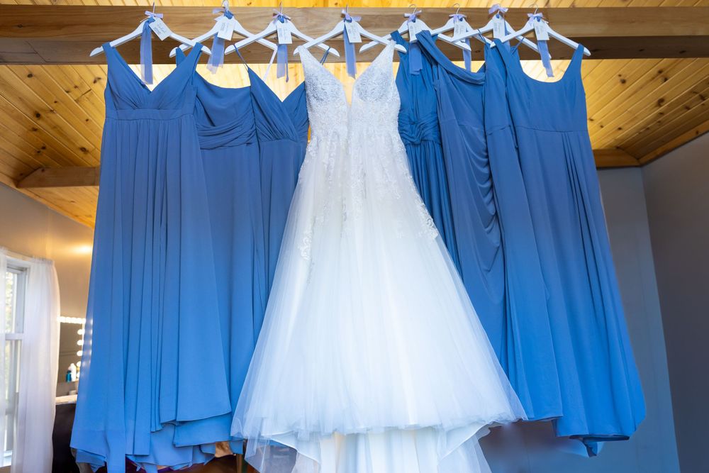 Wedding dress and blue bridesmaid dresses hanging from wooden hangers against wood beam ceiling.
