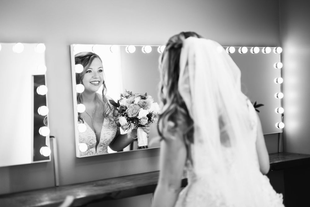 A bride in a white wedding dress and veil looks at her reflection in a makeup room mirror while holding a bouquet.