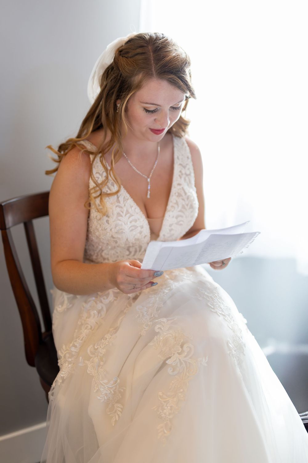 A bride in a lace wedding gown sits in a wooden chair reading a letter in soft natural light.