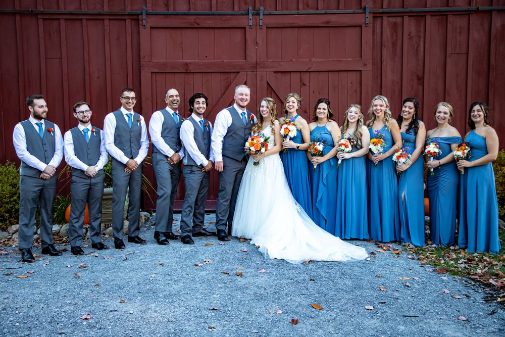 Wedding party poses in front of red barn with bridesmaids in blue dresses and groomsmen in gray vests.