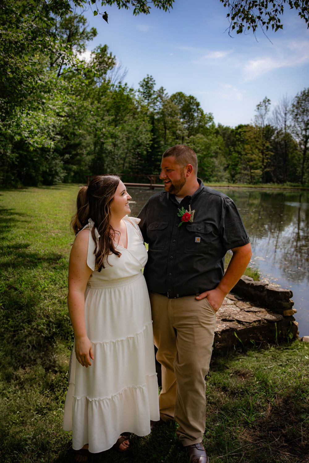 A couple shares a romantic moment by a serene pond surrounded by lush green trees on a sunny day.
