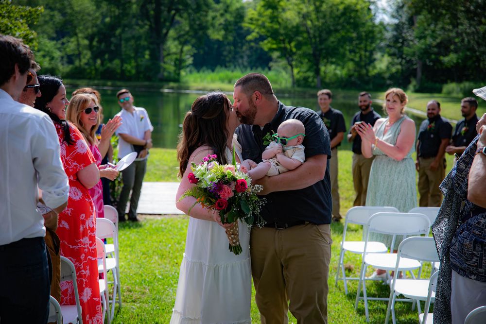 Outdoor wedding ceremony in a grassy field with guests and white chairs on a sunny summer day.