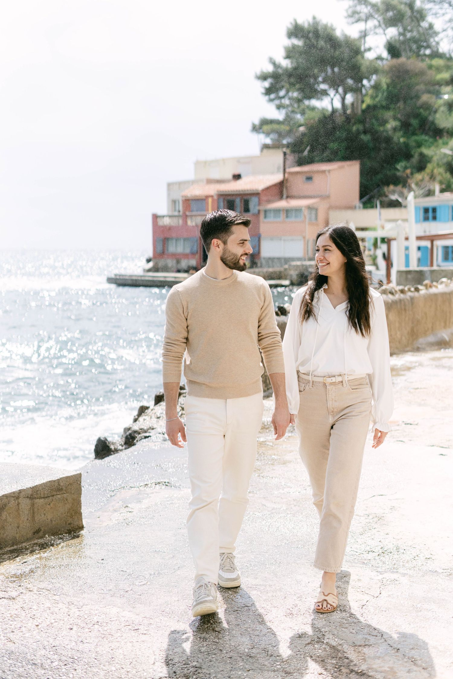 Couple walking together on a coastal path in neutral beige and white outfits with waterfront buildings behind them.