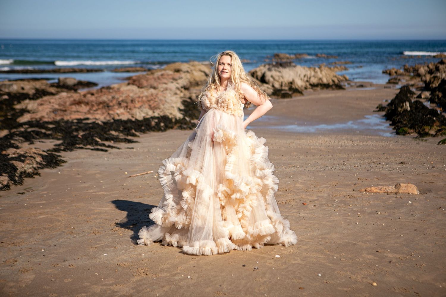 A person in a flowing cream-colored ruffled gown poses gracefully on a beach with rocky outcrops and ocean waves.