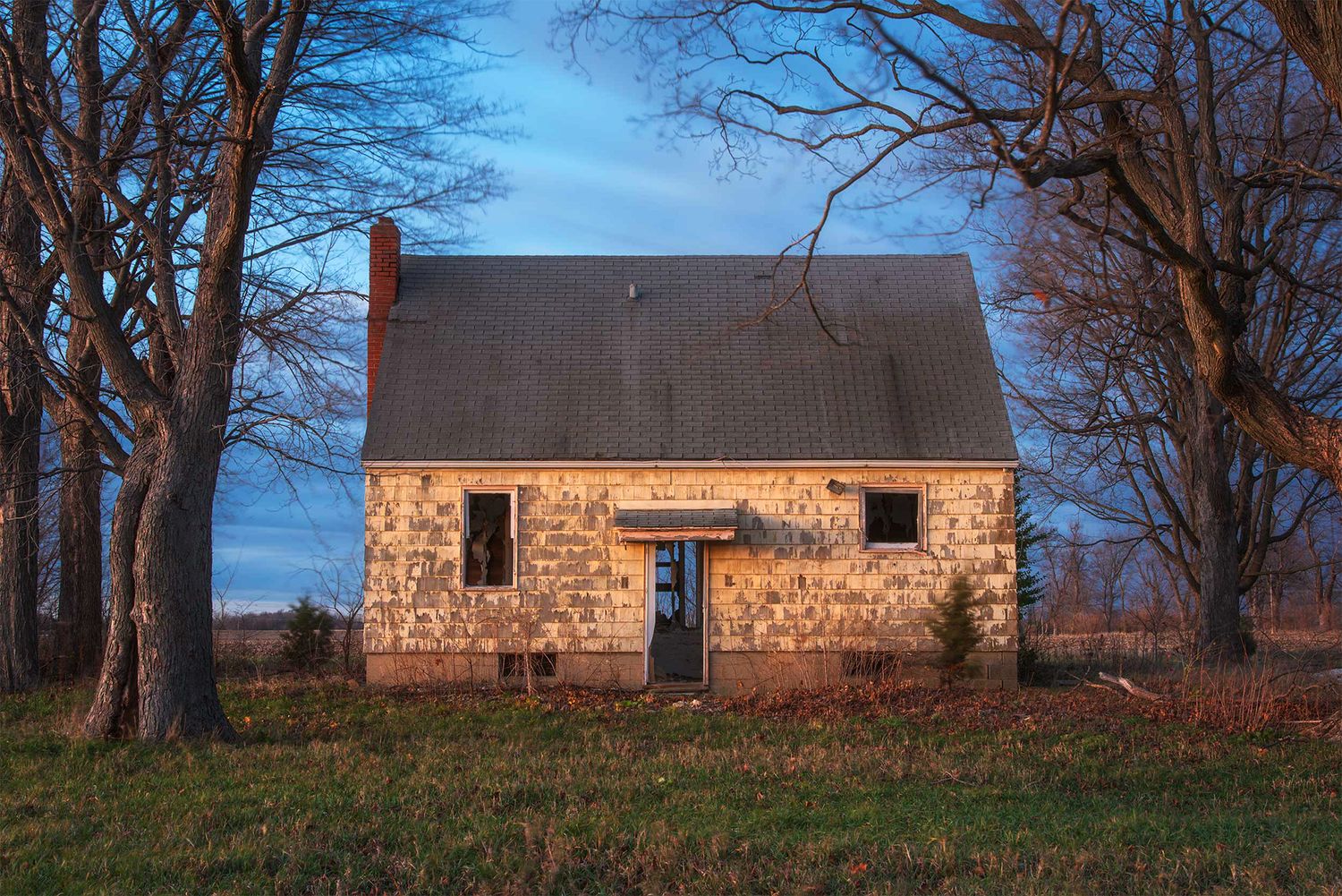 Rustic stone cottage with blue door illuminated at dusk among bare trees.