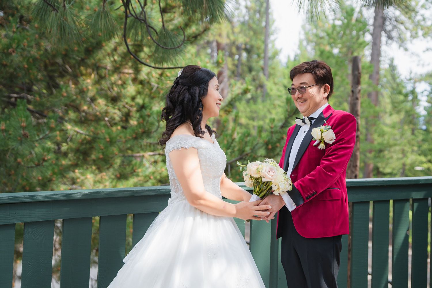 A wedding couple shares a tender moment on a wooden deck surrounded by lush green forest in matching burgundy and white attire.