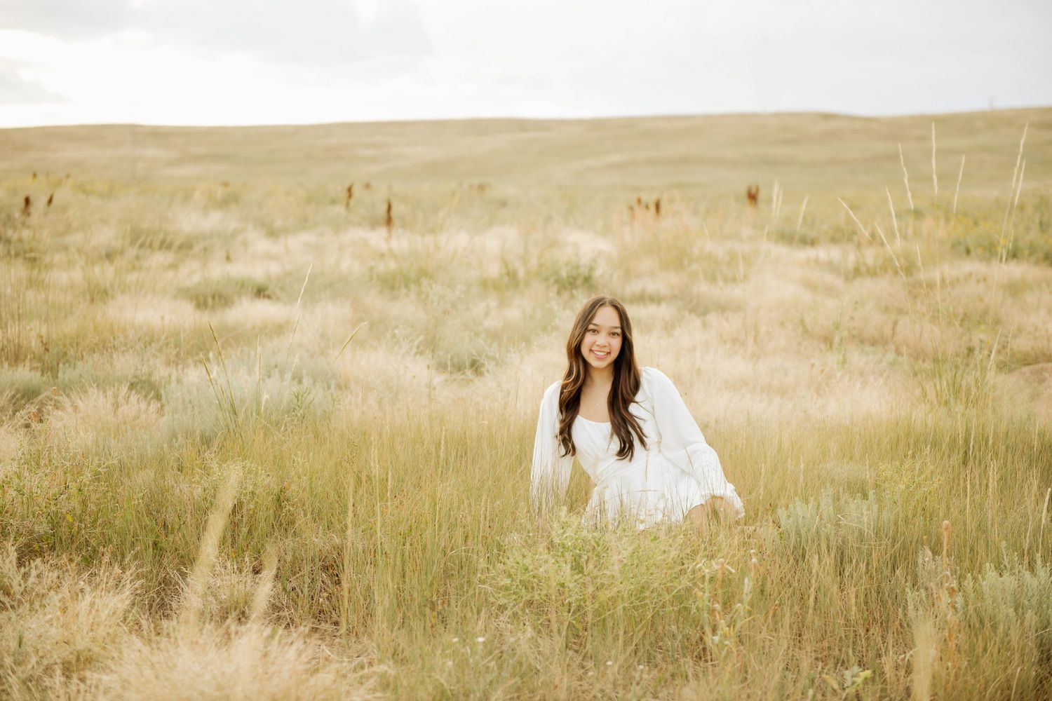 A tranquil beach portrait session in soft natural light showing a woman in a white dress sitting among coastal grasses.