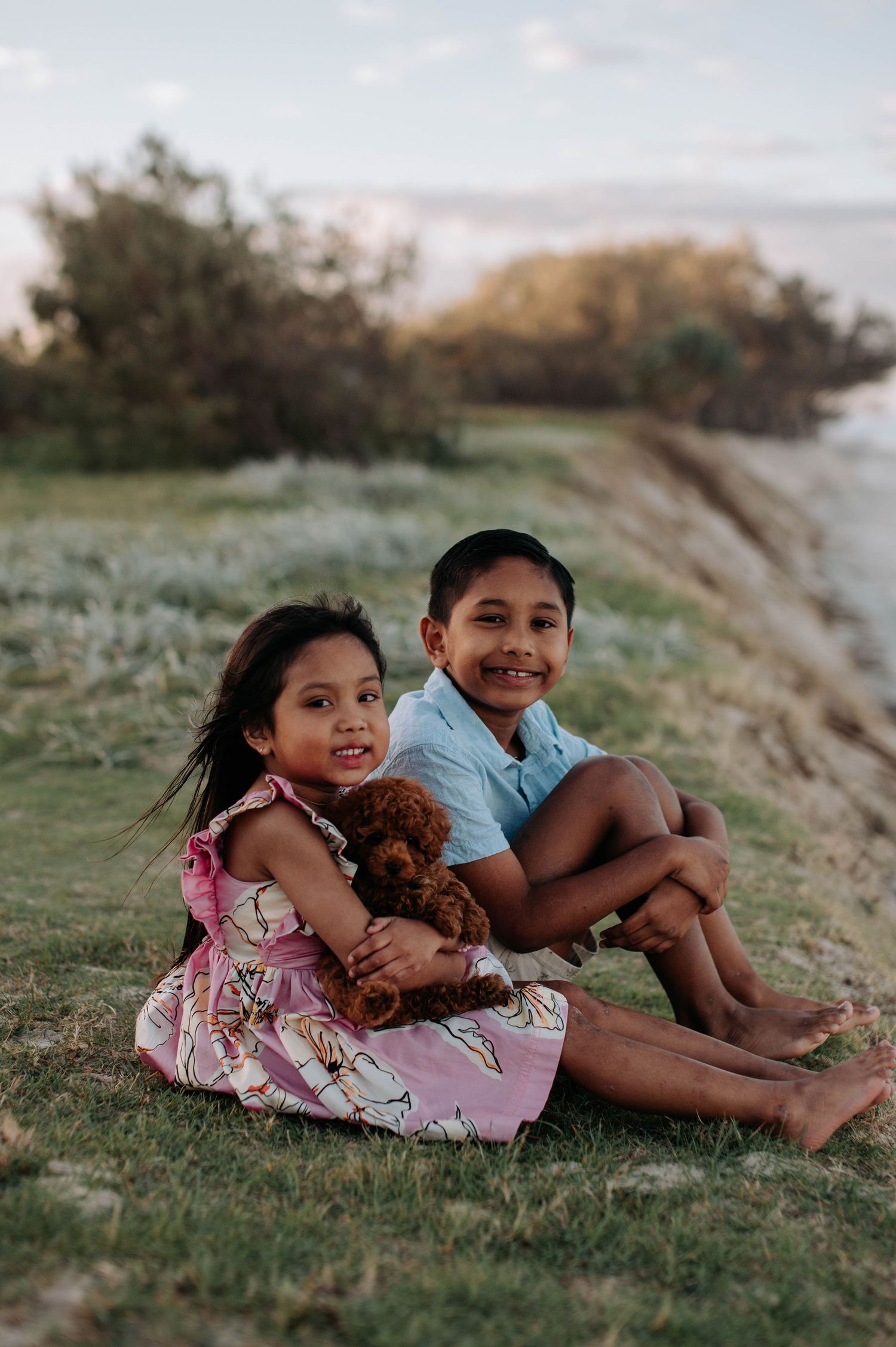 Two children lying in grass with a small brown dog during golden hour lighting.