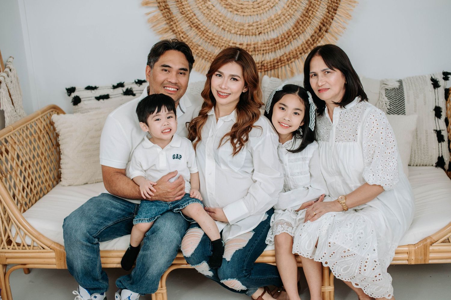 Group wearing white tops and jeans sit together on a cozy woven bench for casual indoor family photos.