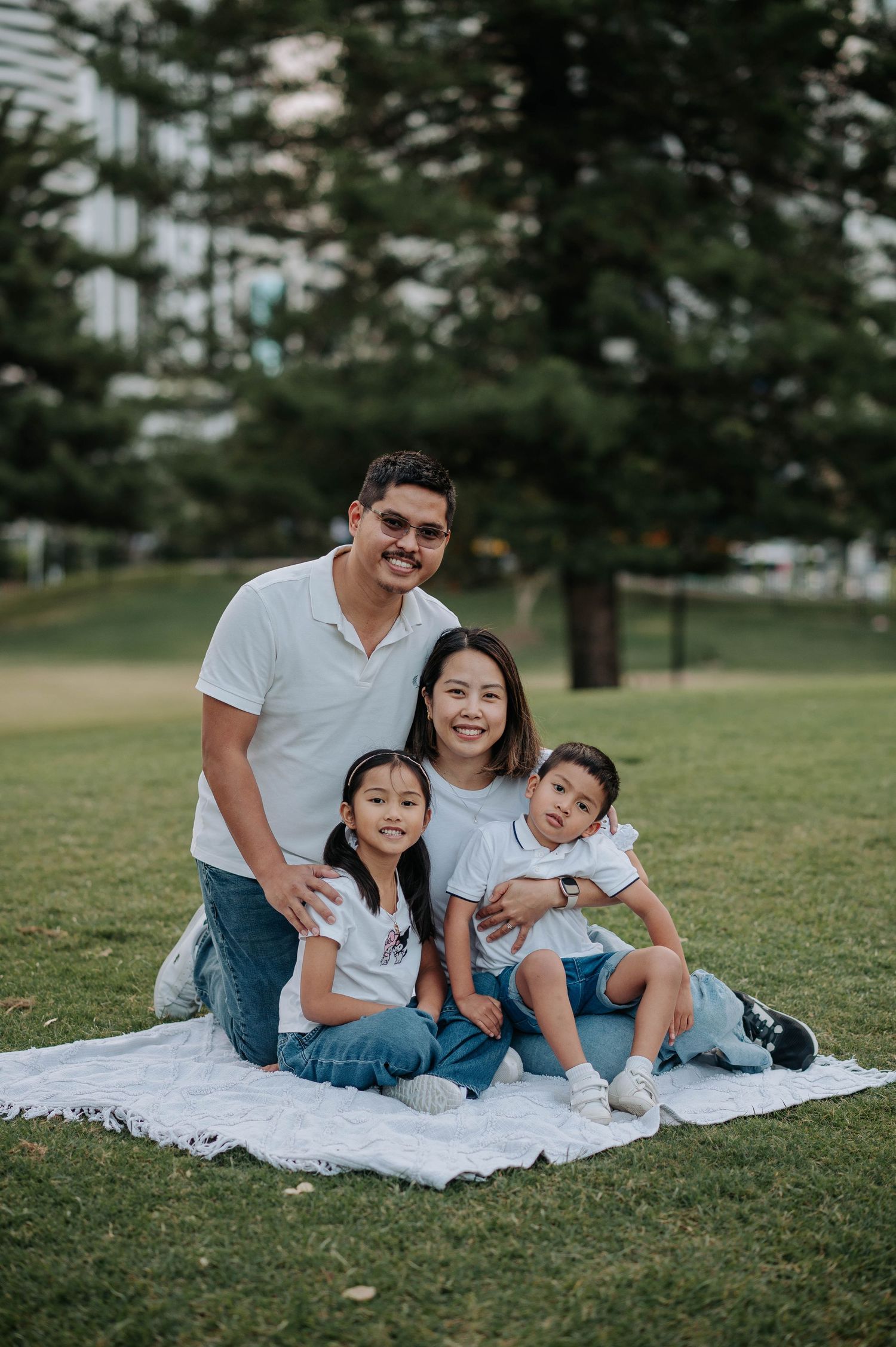 A loving family of four sits together on a white blanket in a grassy park during a relaxed outdoor photoshoot.