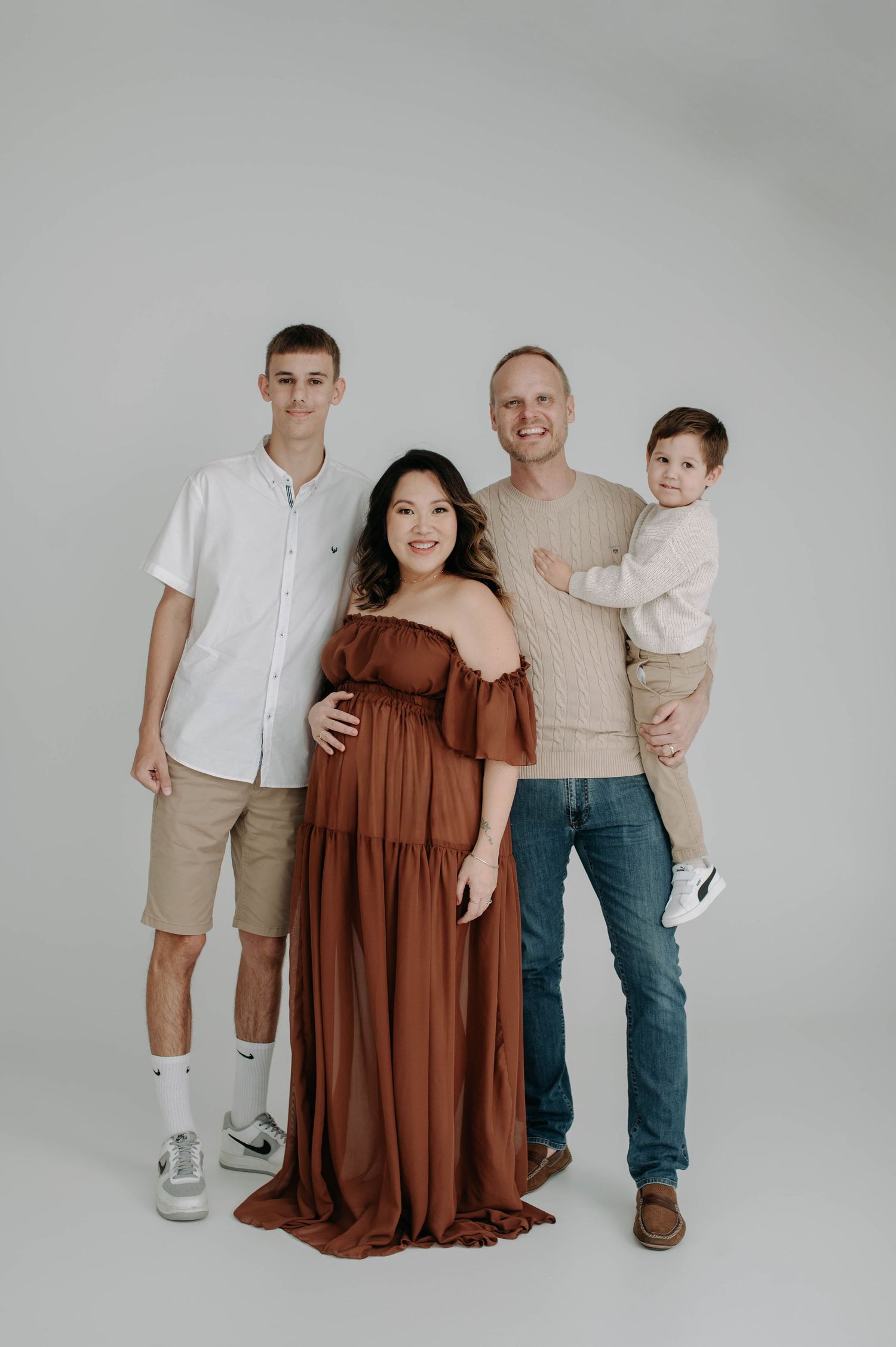 Family portrait studio photo with neutral background showing group in coordinated casual outfits of white and brown tones.