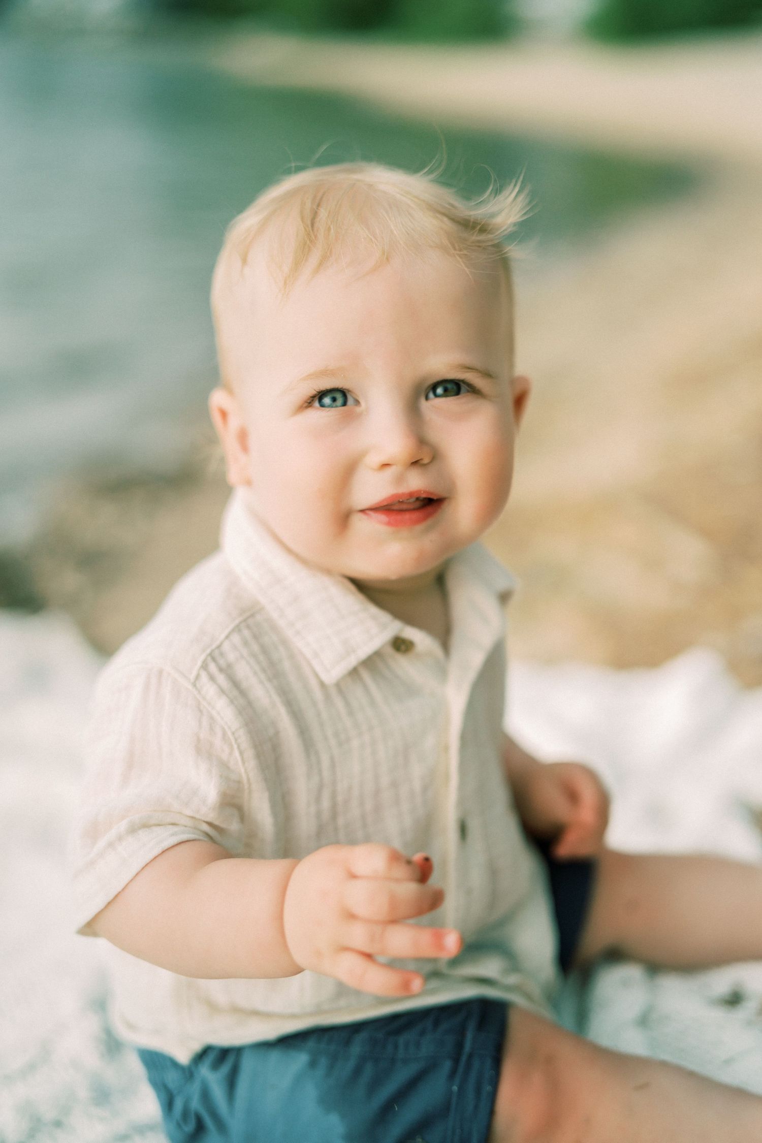 A parent wearing a blue floral dress holds a baby while standing barefoot on sandy ground with greenery in the background.