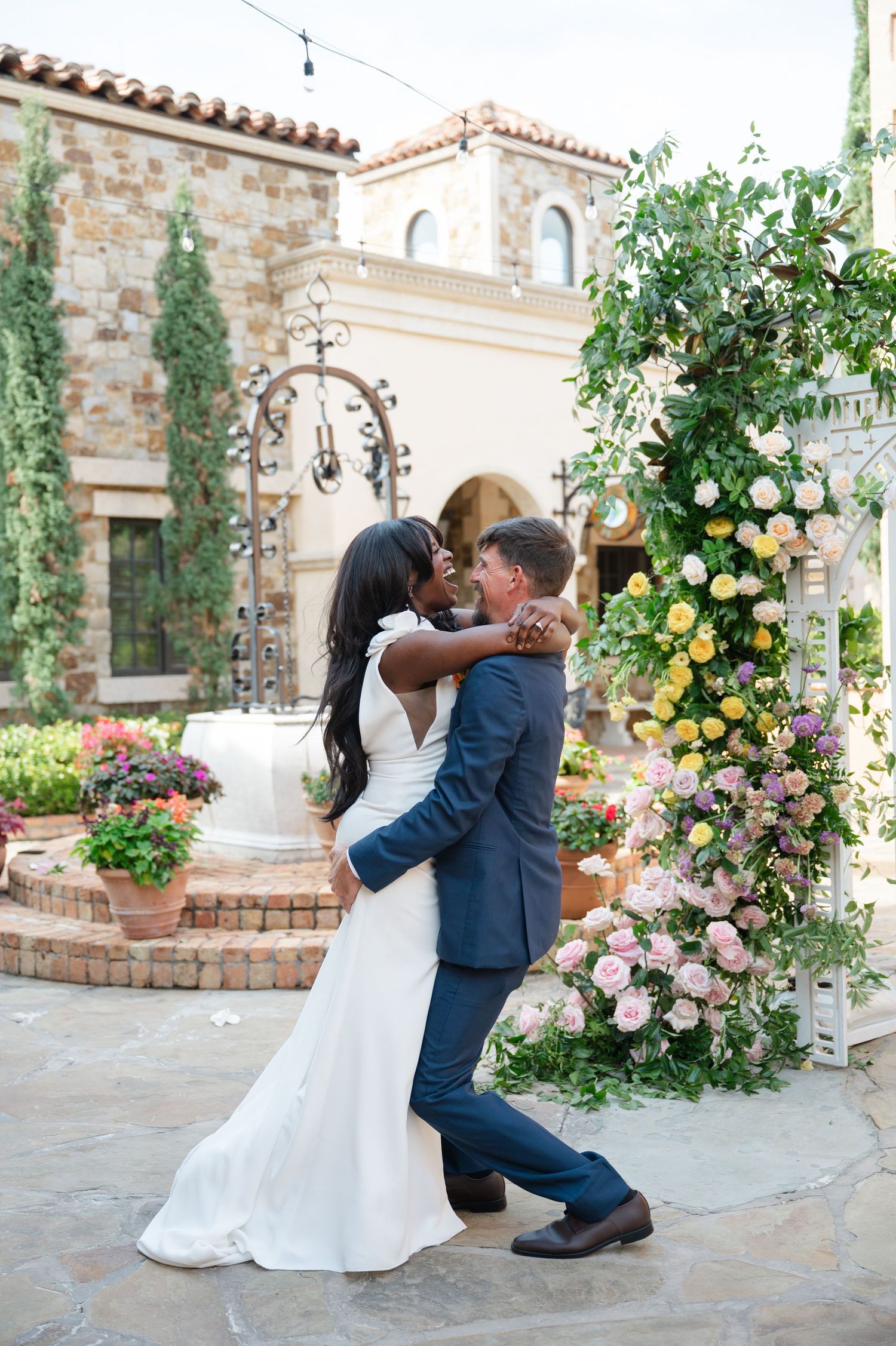 A romantic wedding couple embraces in a garden courtyard with stone walls and blooming roses at a Spanish-style venue in NYC