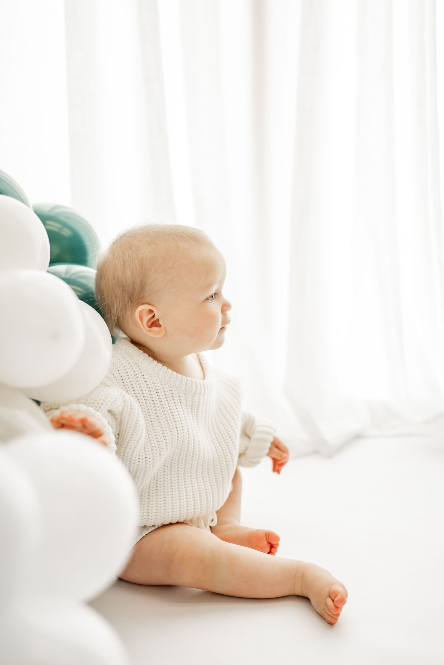 Baby wearing white sweater lying down on soft white fabric in gentle, minimalist nursery setting.