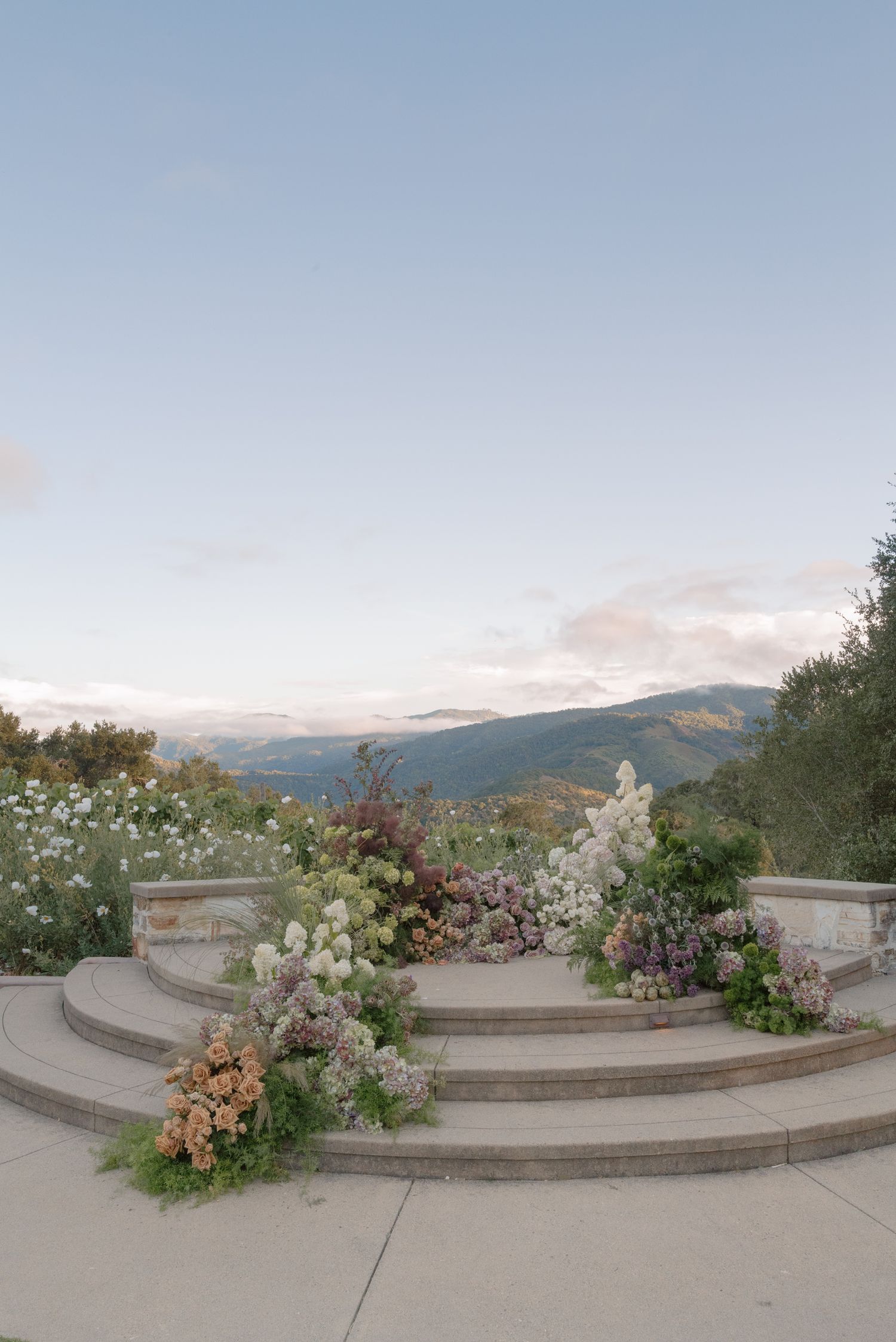 A panoramic view of a curved stone terrace with flowering plants and mountains visible in the background under a cloudy sky.