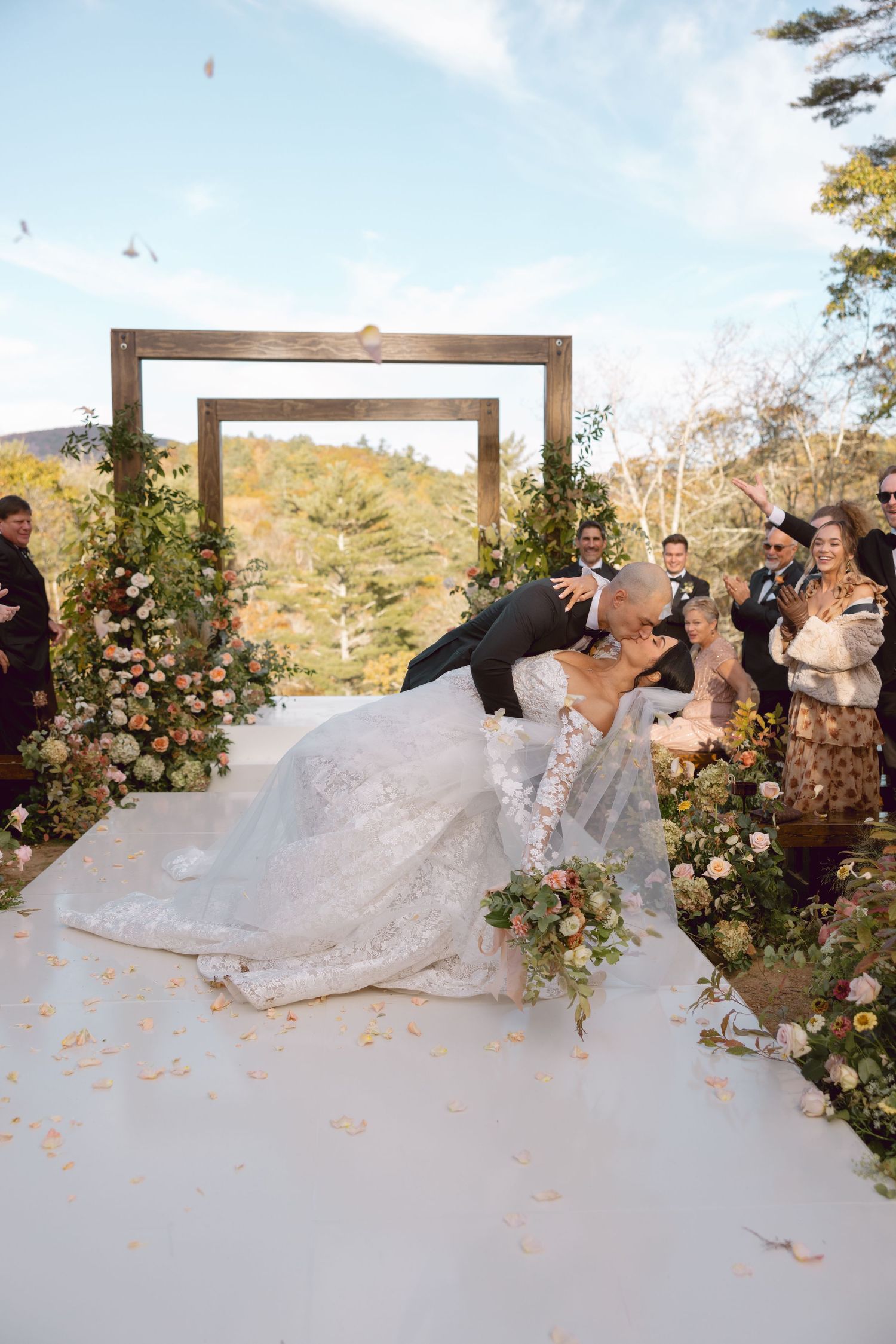 A romantic wedding ceremony dip kiss under a floral arch in an outdoor mountain setting with guests looking on.