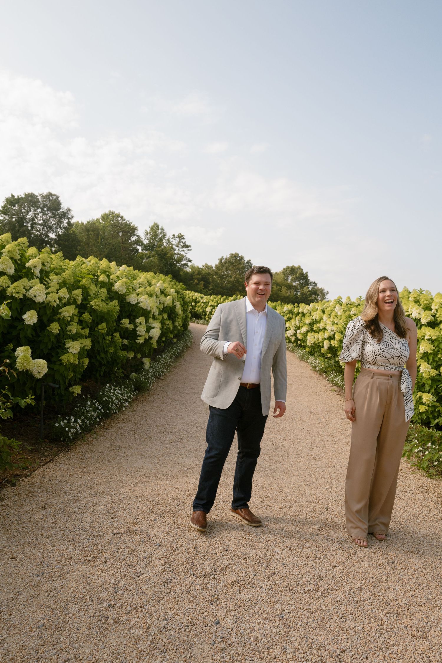 Two people walking down a gravel path lined with lush hydrangea bushes and greenery on a sunny day.