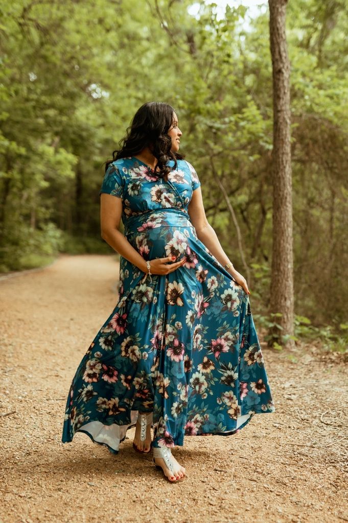 A pregnant woman in a flowing blue floral maxi dress stands on a dirt path in a wooded setting.