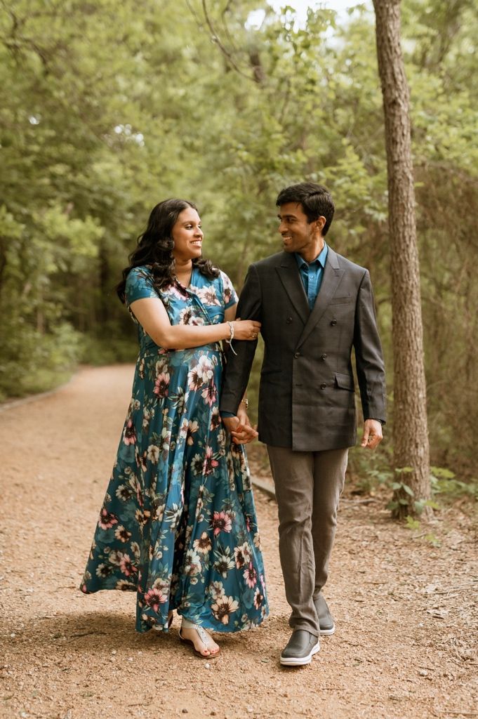 Couple walking together on a dirt path in the woods, wearing formal attire with a floral maxi dress and suit.