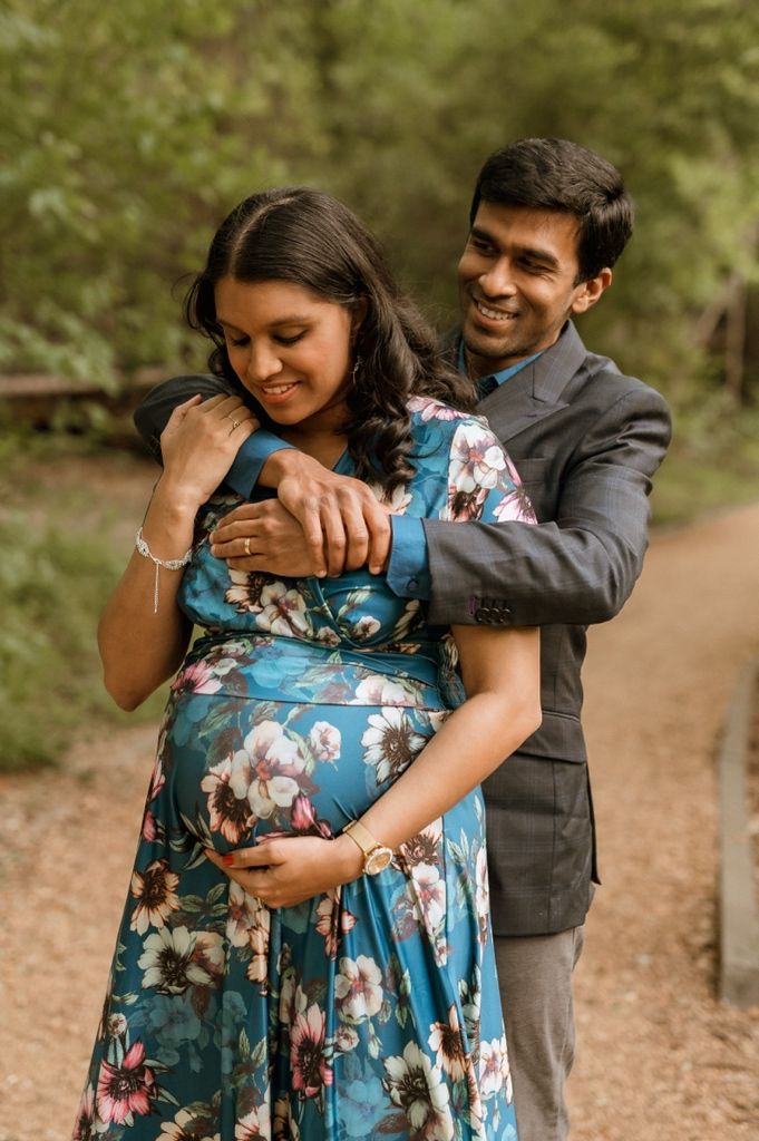 A couple embraces on a woodland path as they celebrate pregnancy in a blue floral maternity dress and gray suit.