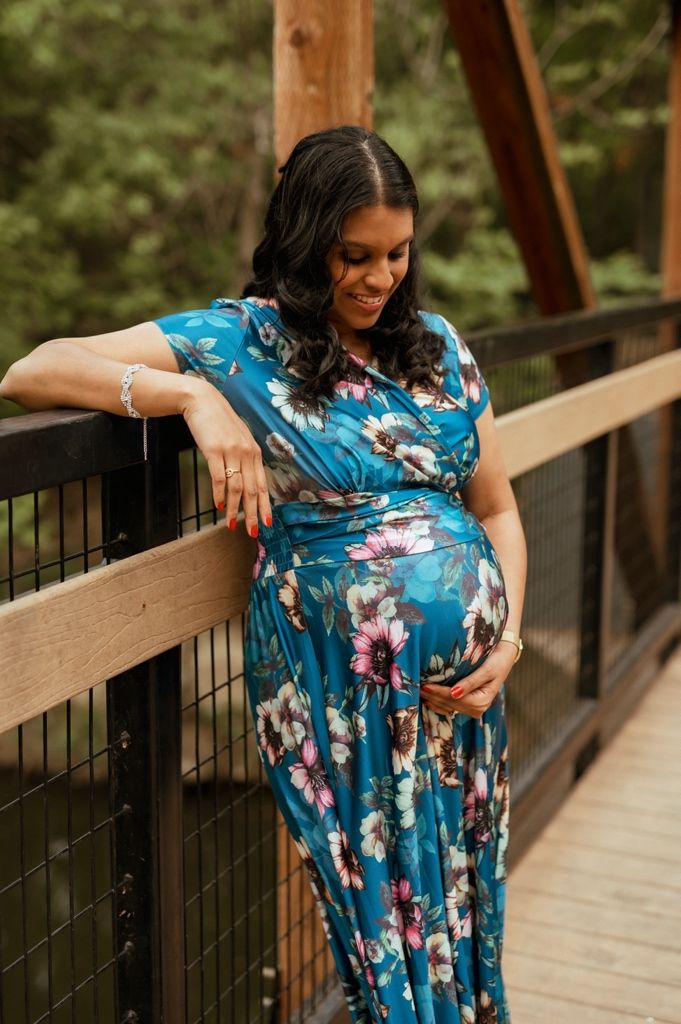 A pregnant woman in a flowing blue floral dress stands on a wooden bridge in a peaceful forest setting.