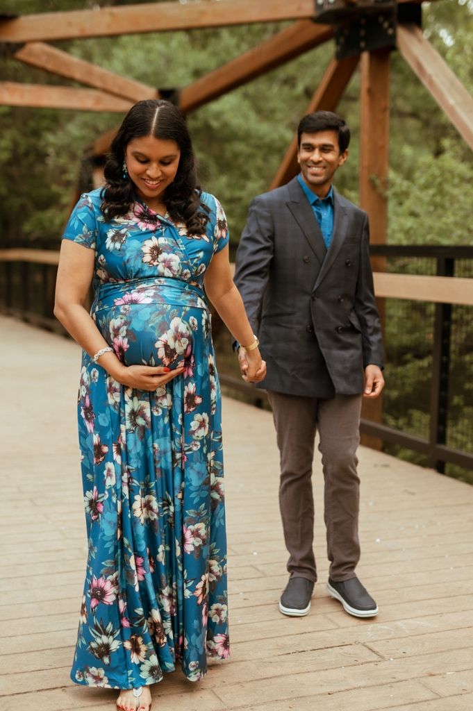 Person in a flowing blue floral dress with pink accents stands on a rustic wooden bridge pathway.