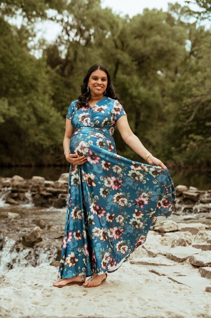 A person in a flowing blue floral dress poses gracefully on rocky terrain surrounded by trees.
