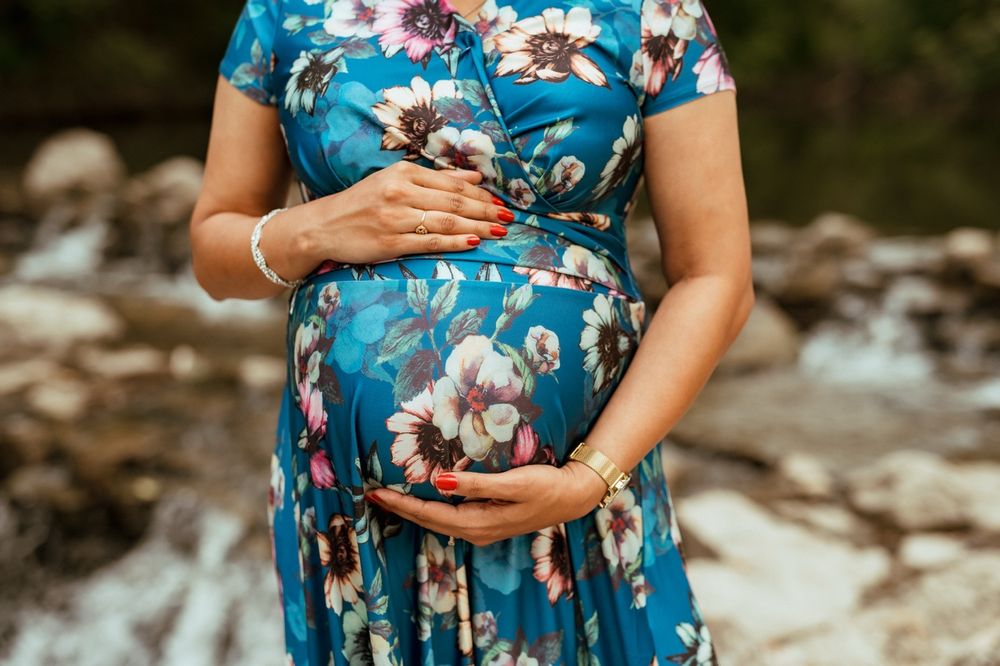 A pregnant woman in a blue floral dress cradles her belly while standing near a rocky stream.