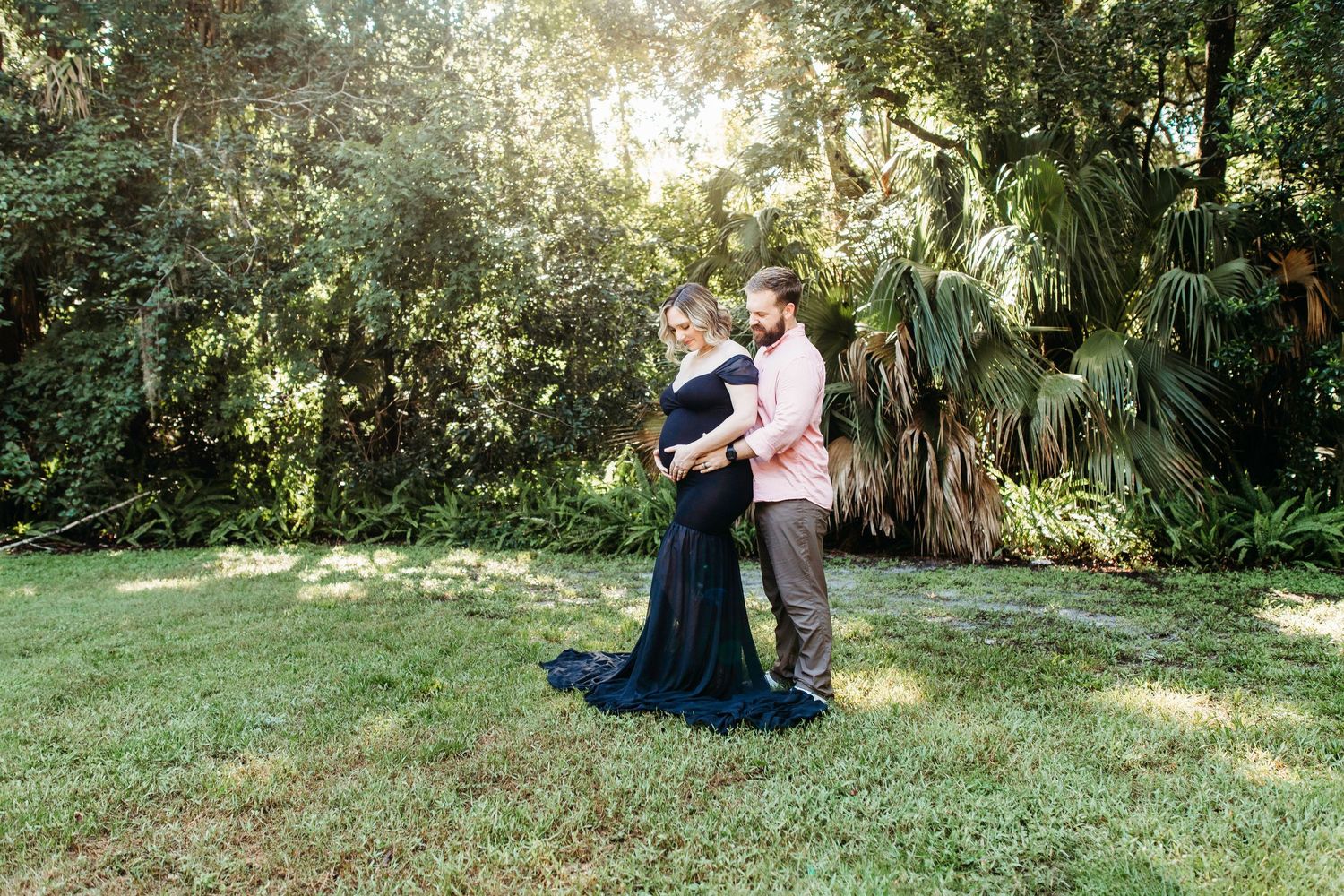 Two people embrace on a sunlit grassy lawn surrounded by tropical foliage and palm trees.