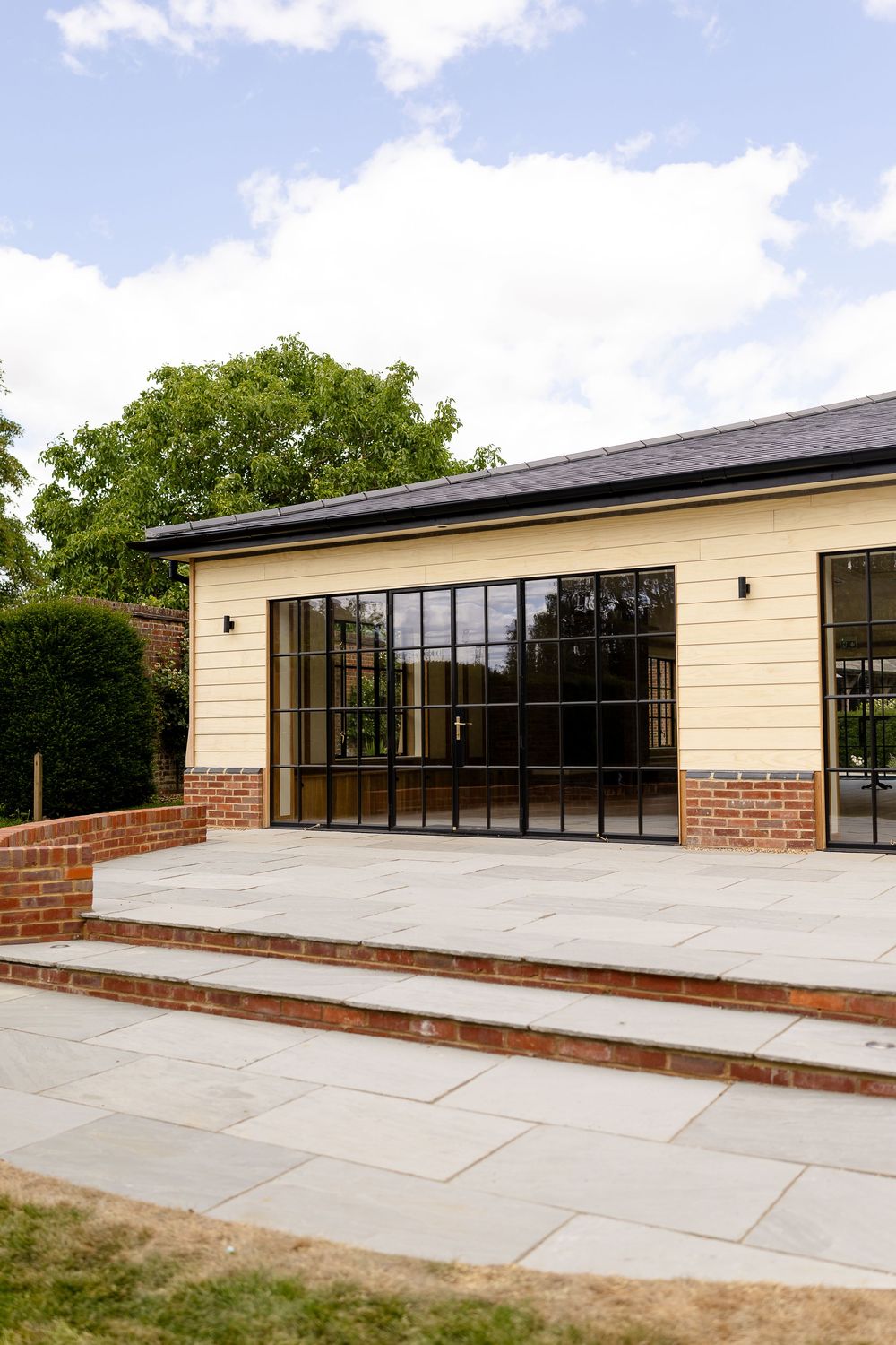 Modern building exterior with glass doors, brick accents, and concrete steps leading to a raised patio area under blue sky.