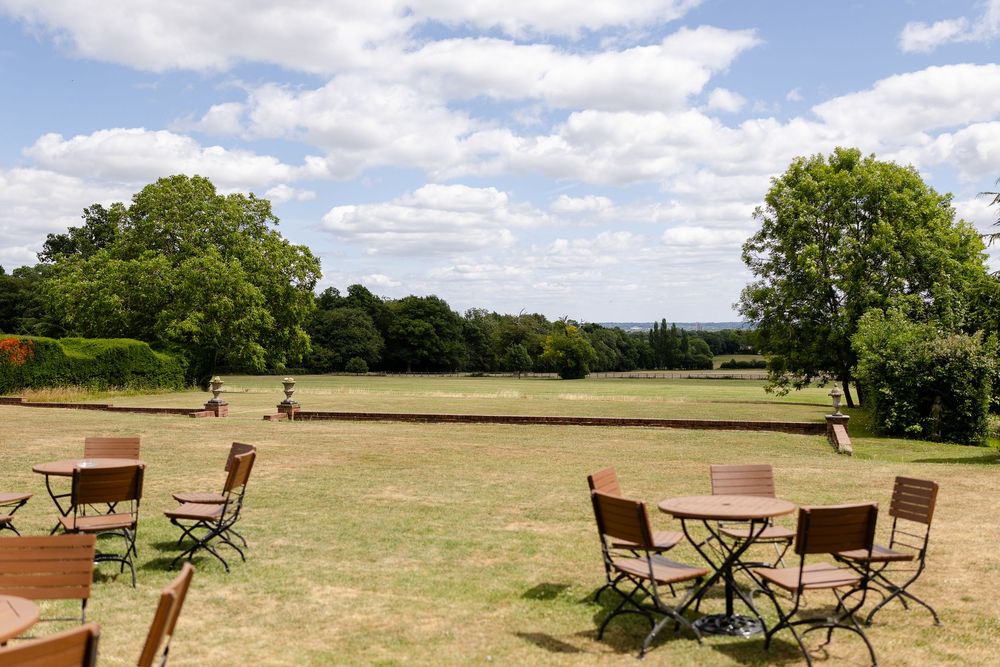 Wooden cafe tables and chairs scattered on a grassy lawn overlooking scenic countryside views on a sunny summer day.