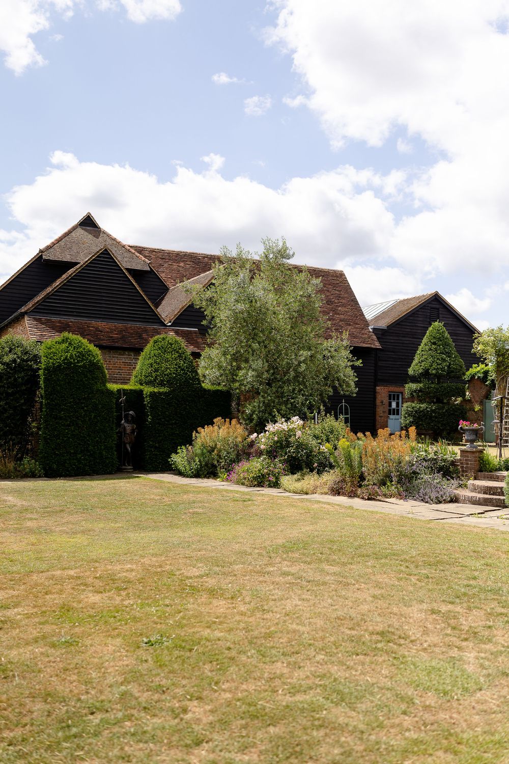 Traditional barn-style country house with manicured topiary gardens and lawn under a bright blue summer sky.