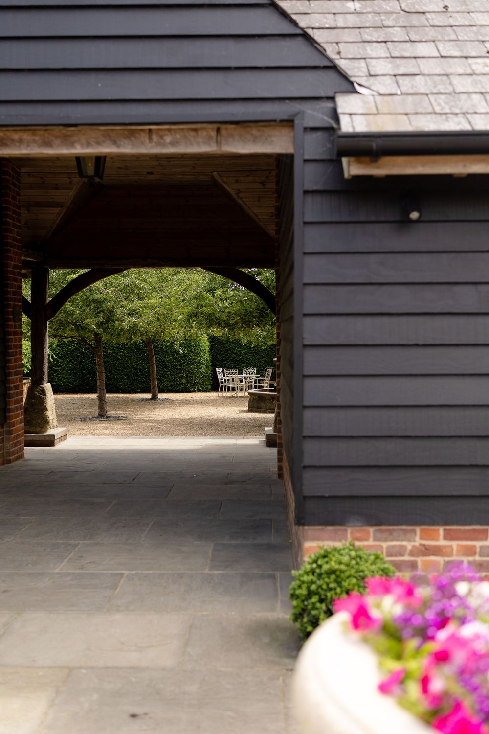 Covered walkway of dark gray building with stone foundation and pink flowers blooming in foreground.