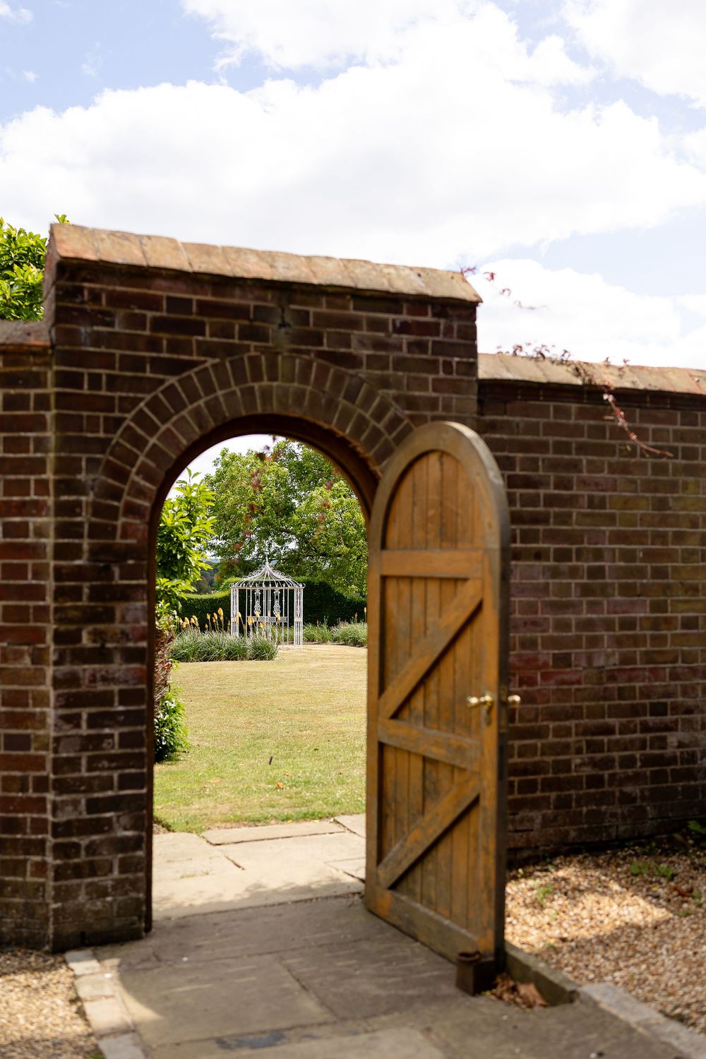 Four arched openings in a brick wall with wooden doors leading to a grassy area on a sunny day.