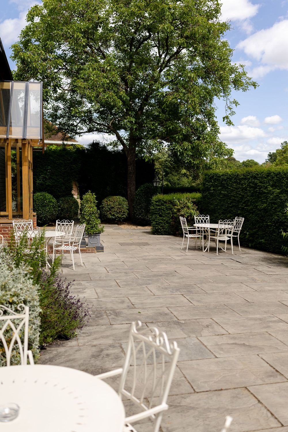 A stone paved outdoor patio terrace with white metal garden furniture and hedges in a sunny garden setting.