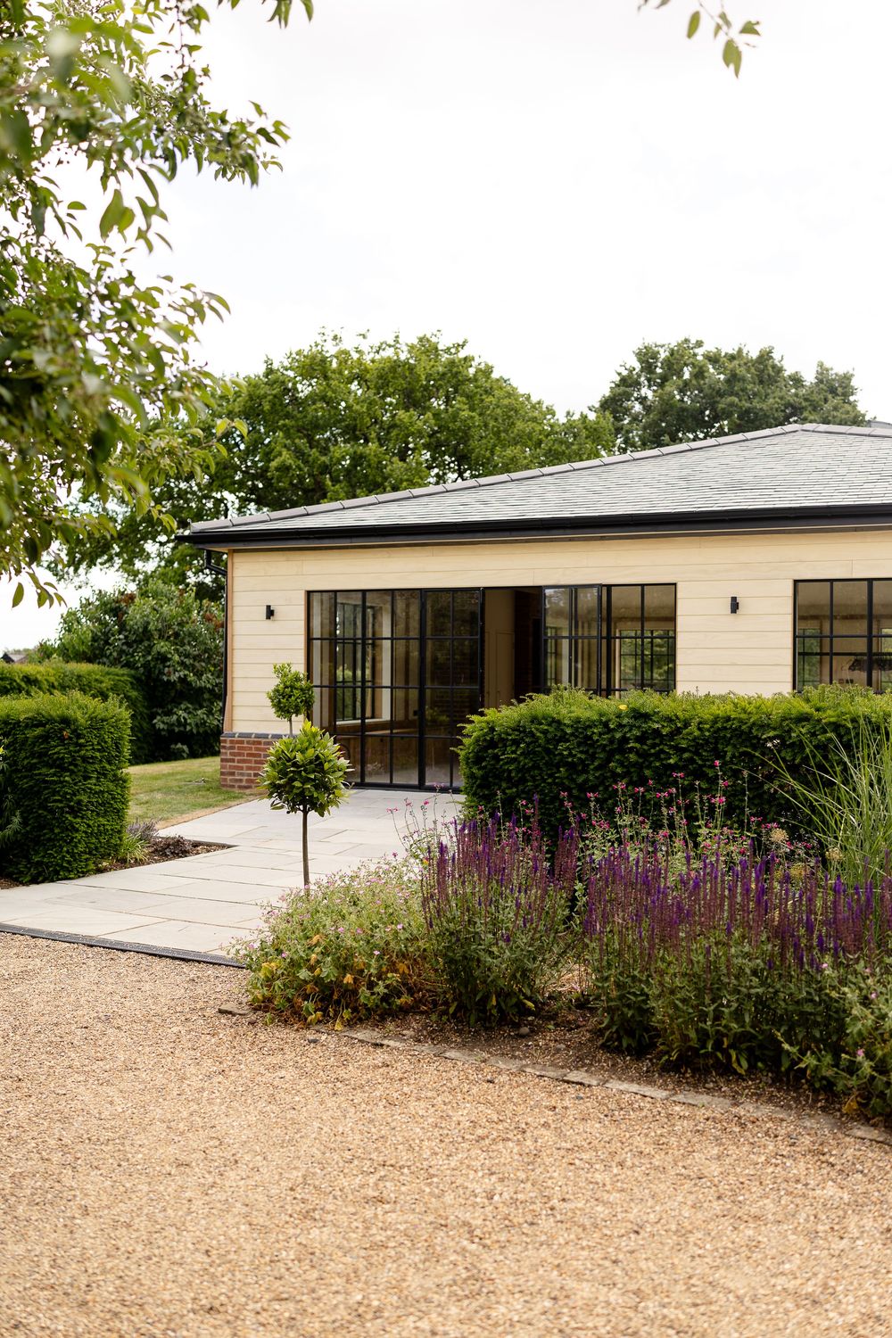 Modern cream-colored house exterior with glass doors, gravel pathway and vibrant purple flowering garden border.