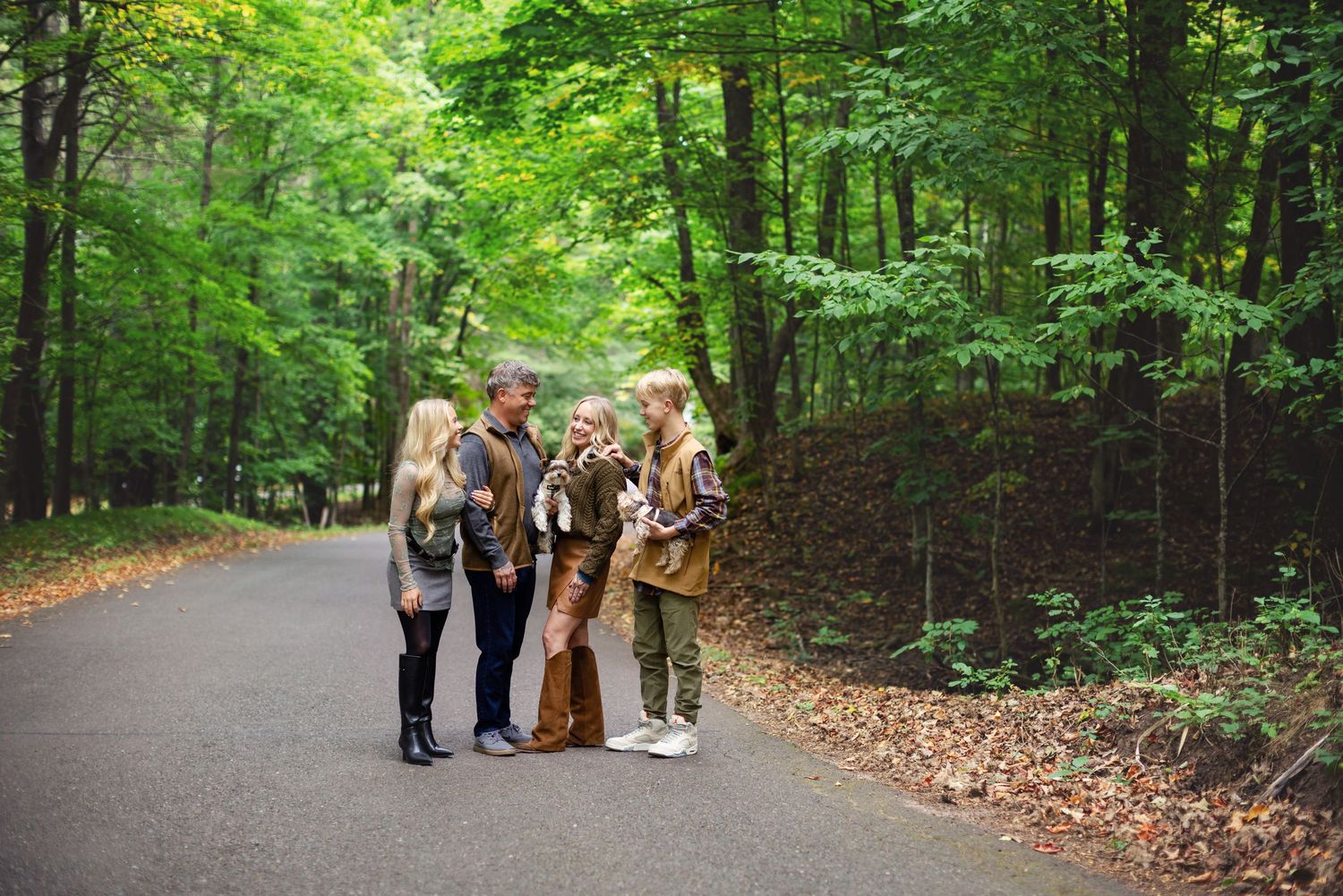 A group walks together along a peaceful wooded path surrounded by lush green trees and fallen leaves.