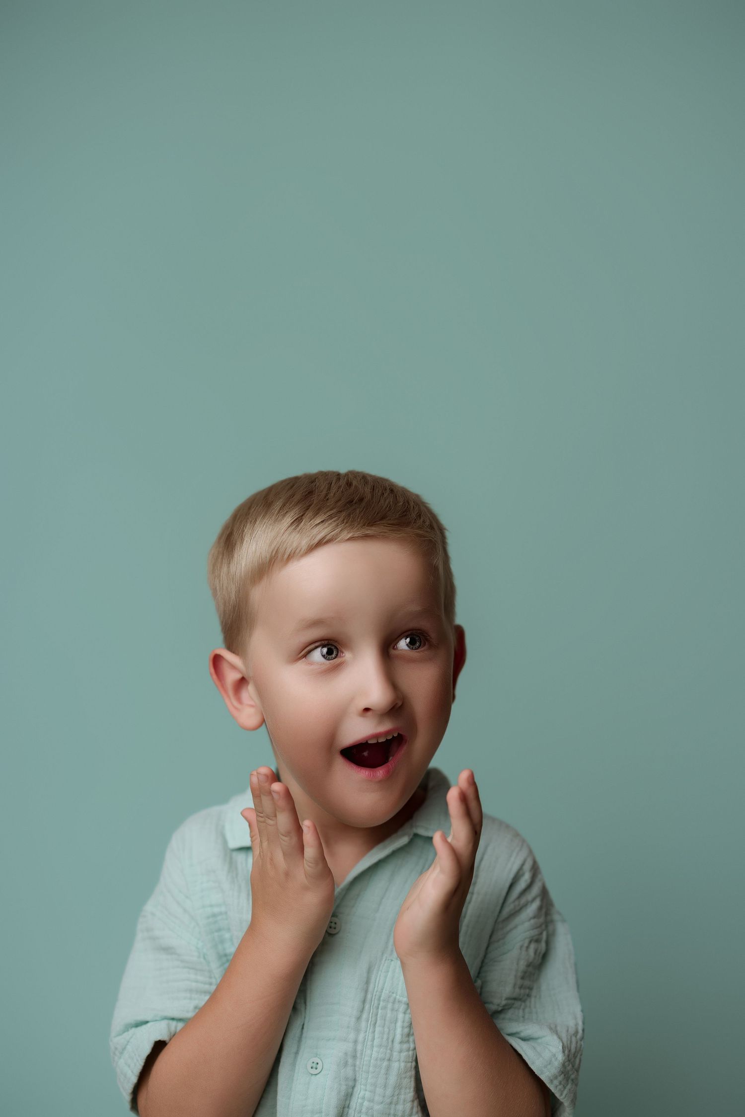 A small child in a mint colored shirt makes silly faces while posing against a teal backdrop in a series of expressions.