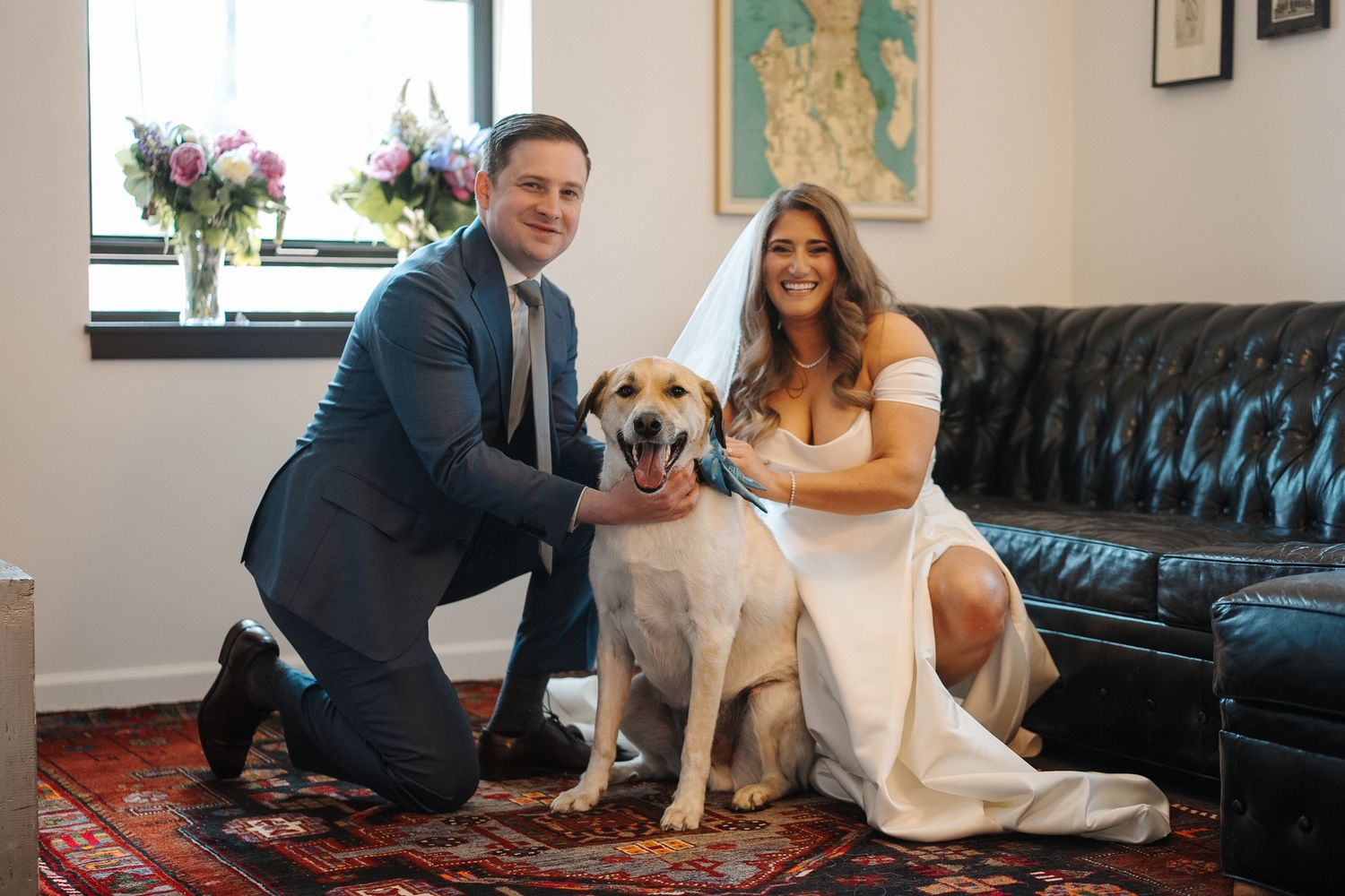 A couple poses with their white and tan dog on a red rug next to a black leather couch.