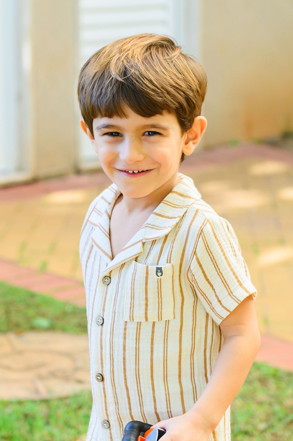 Smiling child wearing a cream colored striped button-up shirt with short sleeves standing outdoors on a sunny day.