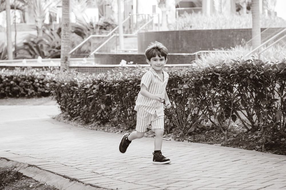 A child in vintage baseball uniform and cleats runs on a brick path next to a manicured hedge in black and white.