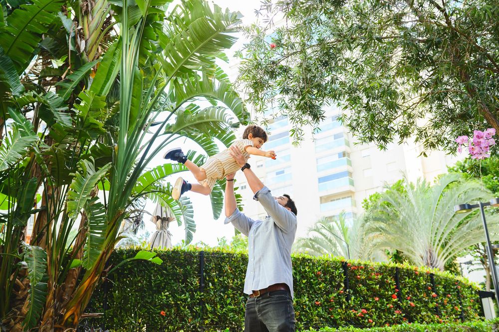 A parent lifts a young child playfully in a lush tropical garden setting with tall palm trees and greenery.