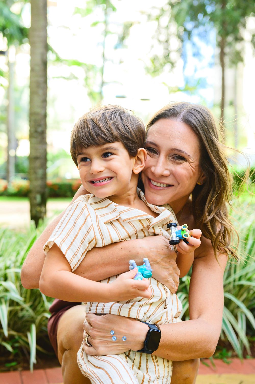 Parent and child share a joyful embrace outdoors while holding small toys in a lush garden setting.